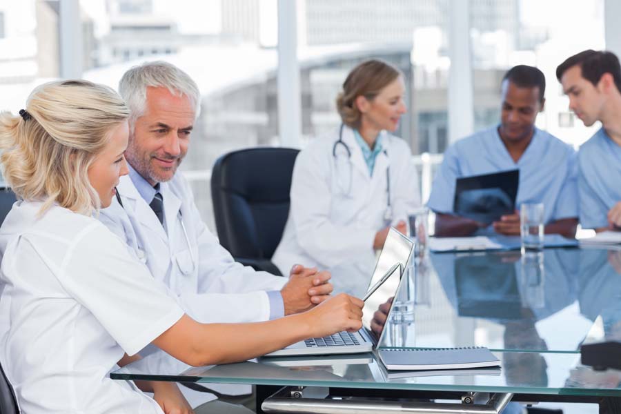 Two doctors look at a laptop together, sitting at a table with other medical professionals.