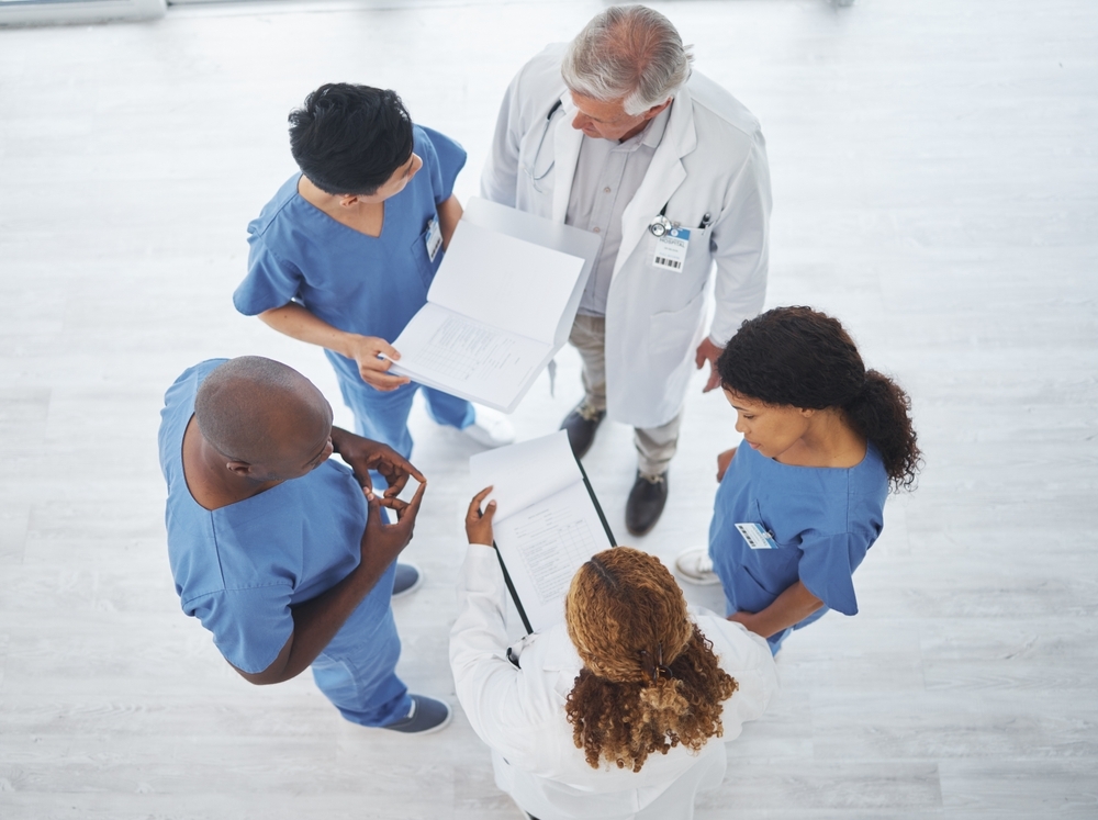 A group of doctors stands in a circle, reviewing documents together. The photo is taken from above.