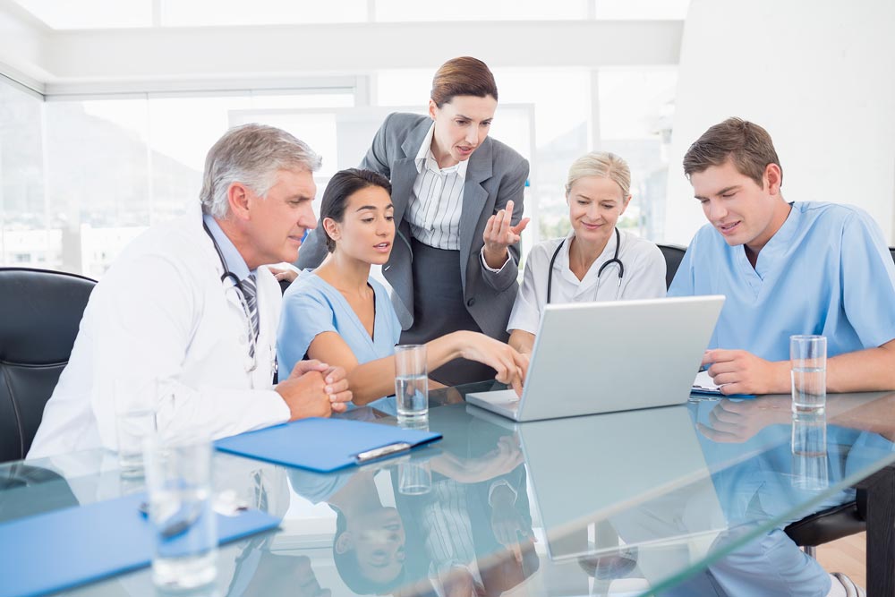 A group of medical professionals sits at a table together, all looking at something on a laptop.